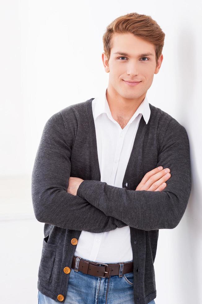 Casually handsome. Handsome young man keeping arms crossed and smiling at camera while leaning at the wall photo