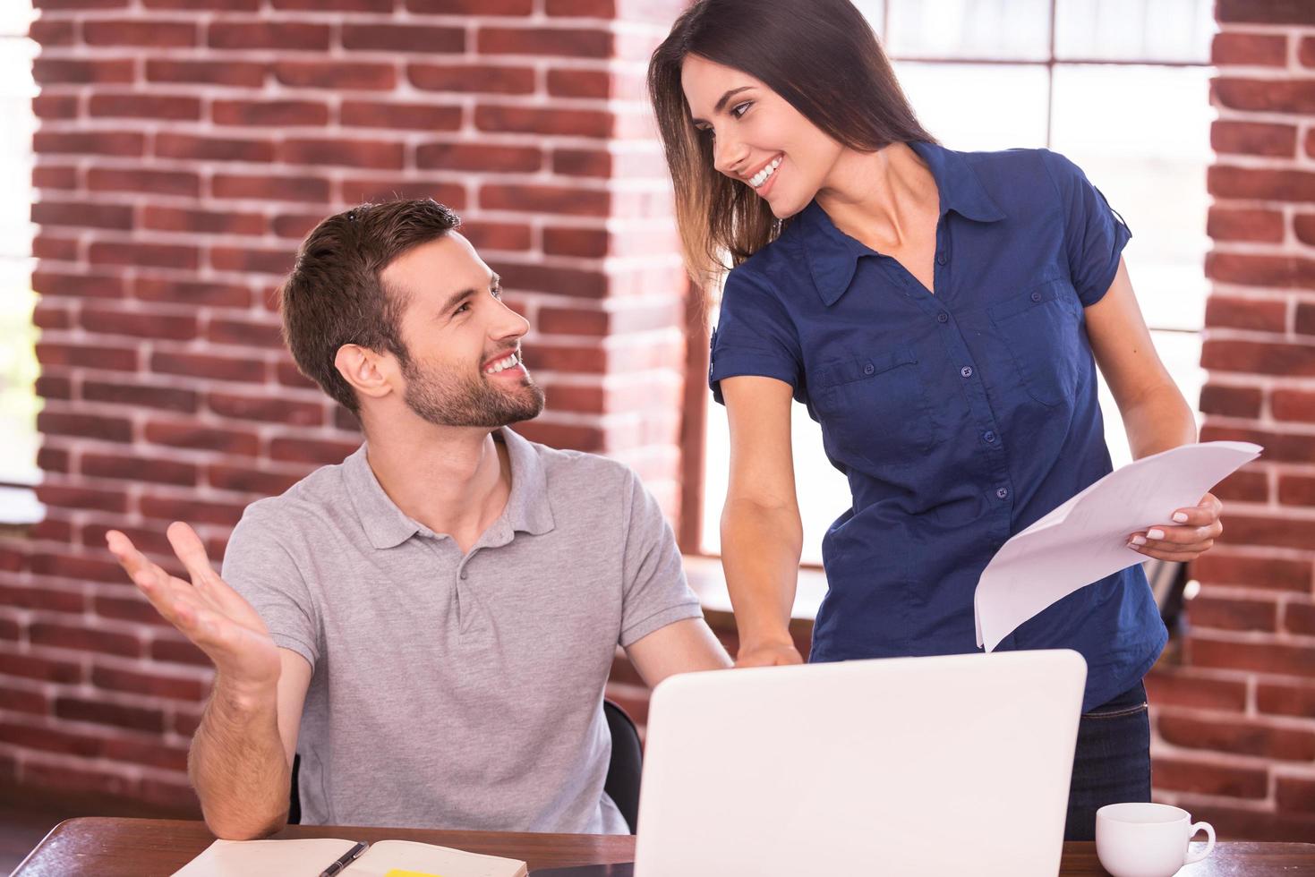 Creative collaboration. Cheerful young man sitting at his working place and gesturing while beautiful woman standing close to him and smiling photo