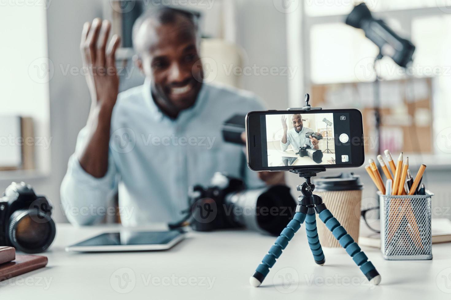 Charming young African man in shirt showing digital camera and telling something while making social media video photo