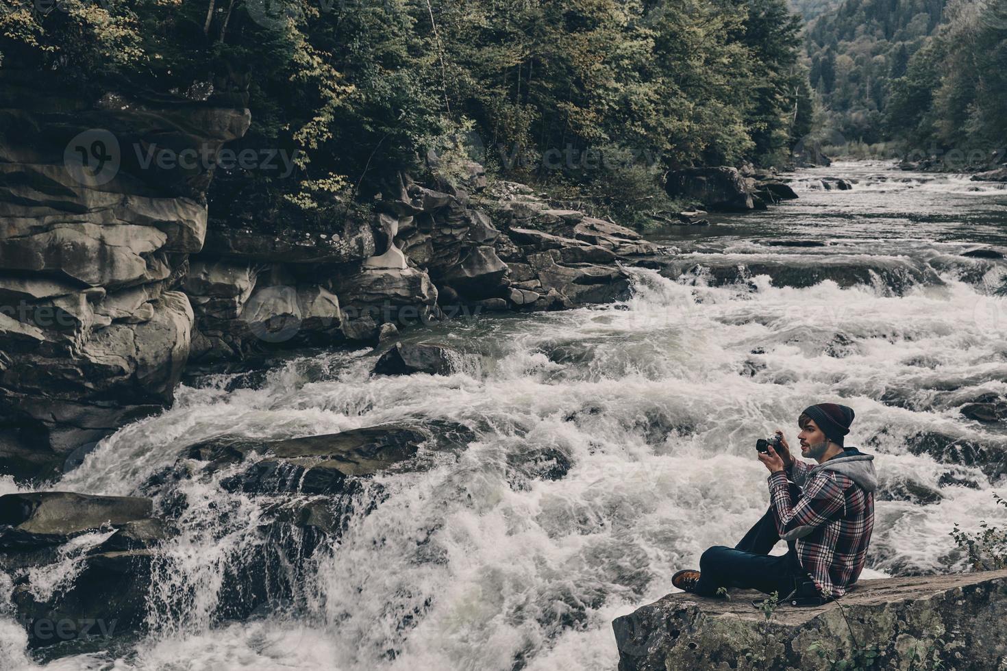 Snapping memories. Handsome young modern man photographing while sitting on the rock near the river photo