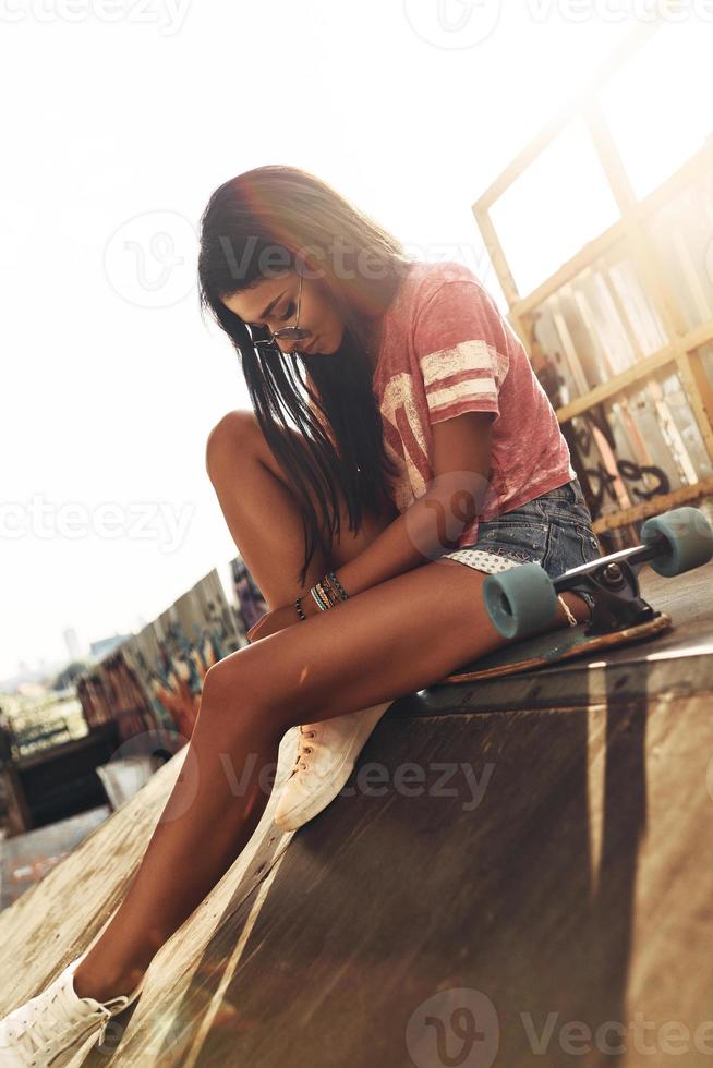 Time to rest. Attractive young woman sitting on the skateboard while spending time at the skate park outdoors photo