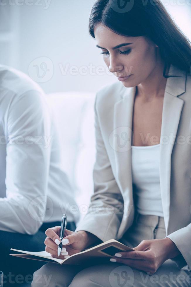 Making some important notes. Close-up part of beautiful young businesswoman writing in notebook while sitting at office with her coworker photo