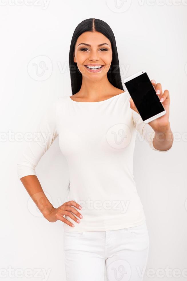 Smart phone for smart people. Beautiful young woman stretching out mobile phone and smiling while standing against white background photo