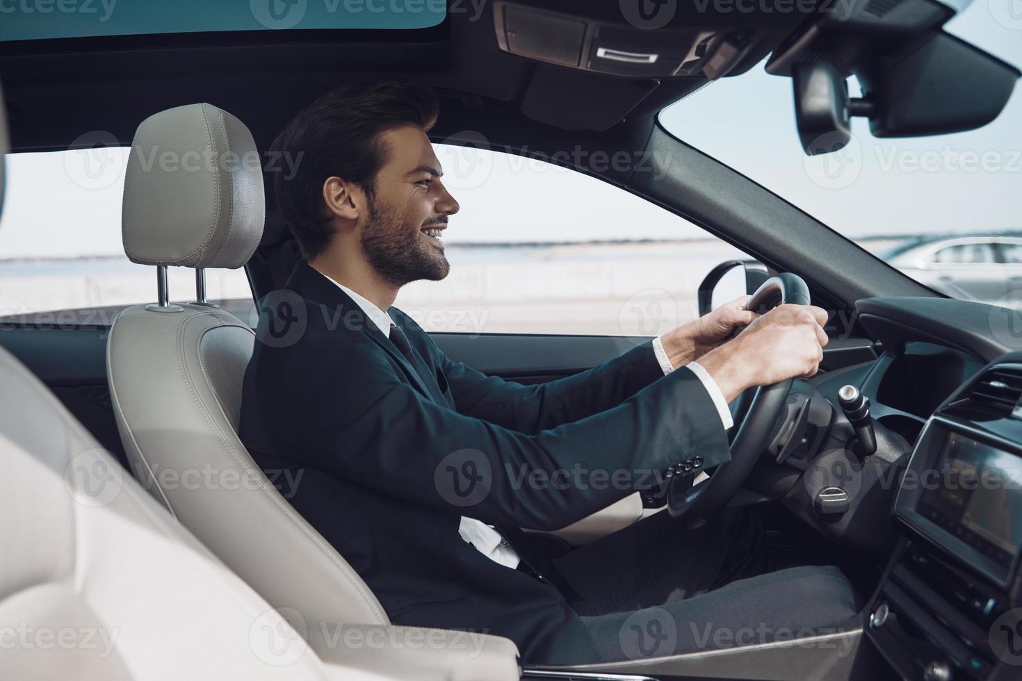 In love with high speed. Handsome young man in full suit smiling while driving a car photo