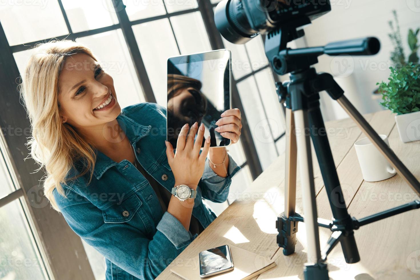 bella joven con ropa informal apuntando a una tableta y sonriendo mientras se sienta frente a una cámara digital foto