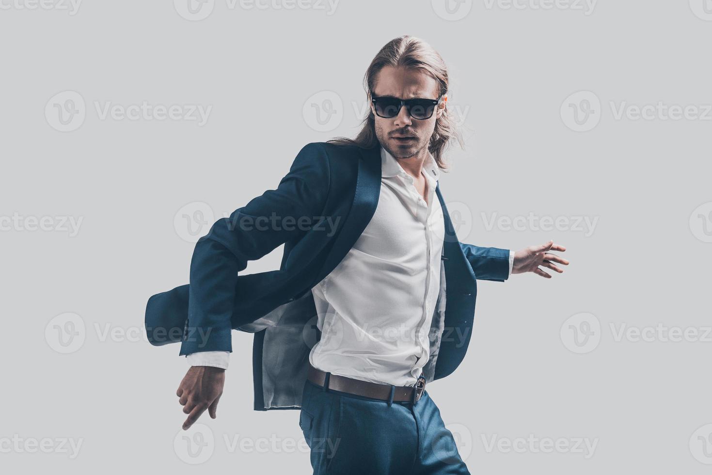 His perfect style in motion. Handsome young man in full suit and sunglasses moving in front of grey background photo