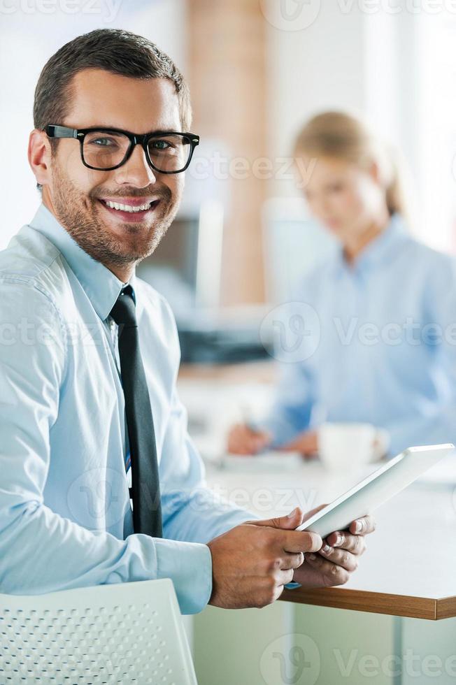 Confident business experts. Cheerful young businessman in formalwear holding digital tablet and looking at camera while his female colleague working in the background photo