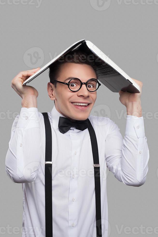 Computer geek. Portrait of young geek in bow tie and suspenders holding a laptop on his head and smiling while standing against grey background photo