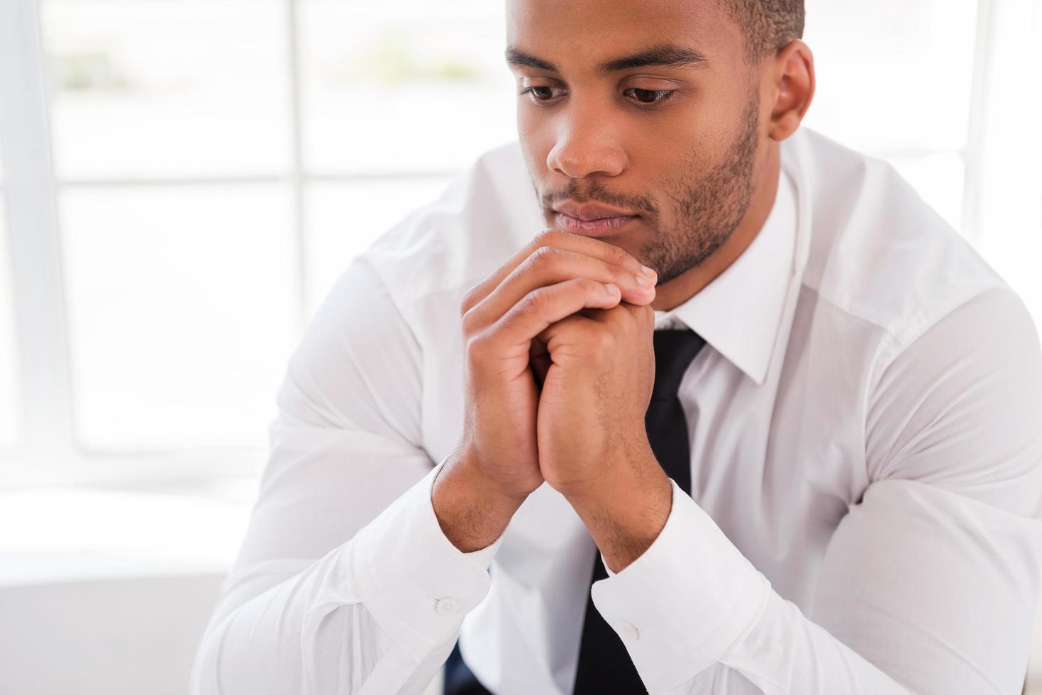 Lost in thoughts. Thoughtful young African man in shirt and tie holding hands on chin and looking away while sitting at his working place photo