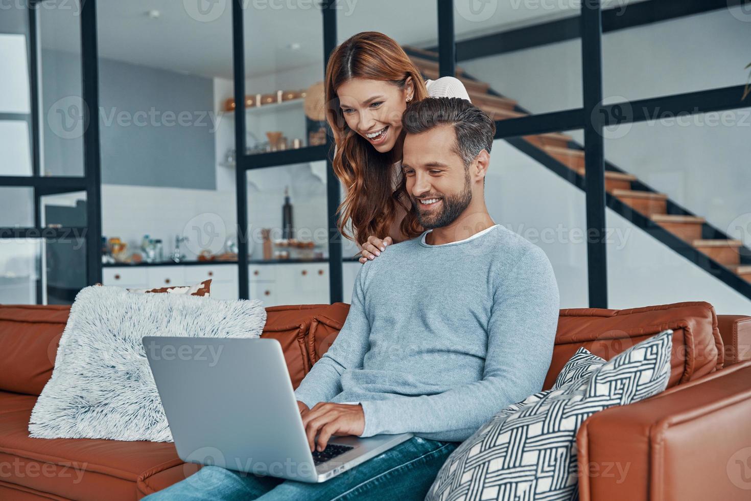 Cheerful young couple using laptop and smiling while spending time at home photo