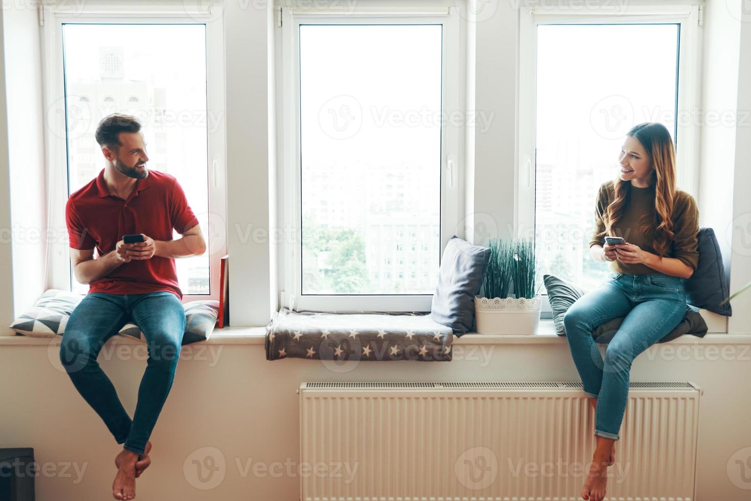 Beautiful young couple in casual clothing holding their smart phones and looking at each other while sitting on the window sill photo