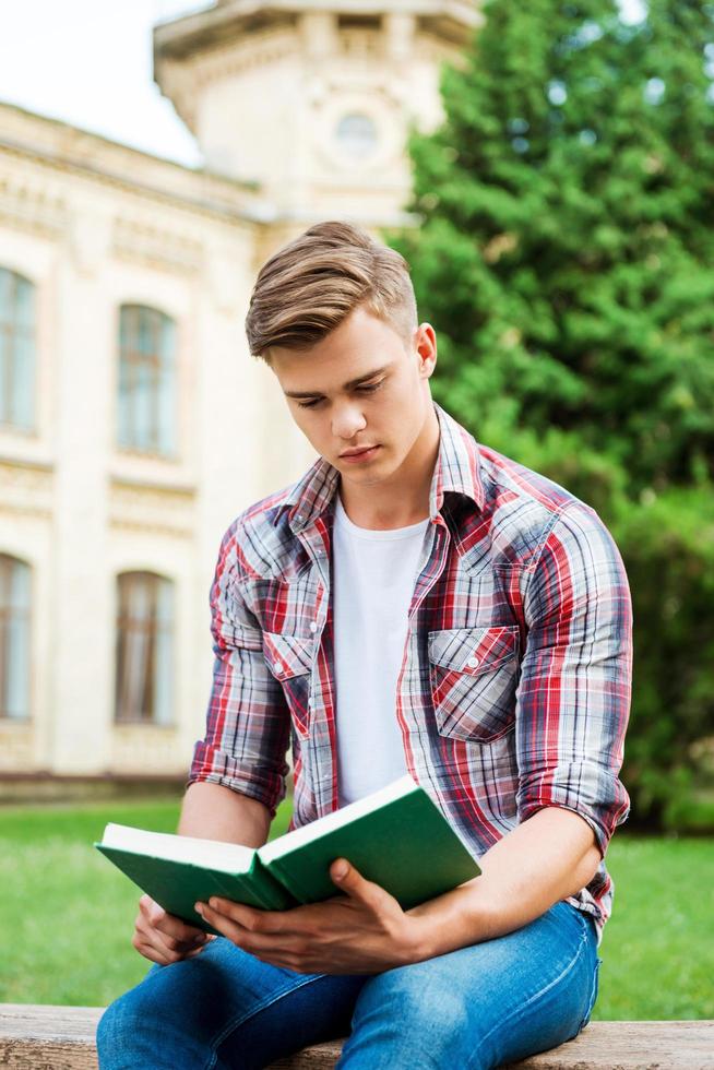 Handsome bookworm. Confident male student reading book while sitting on the bench and in front of university building photo
