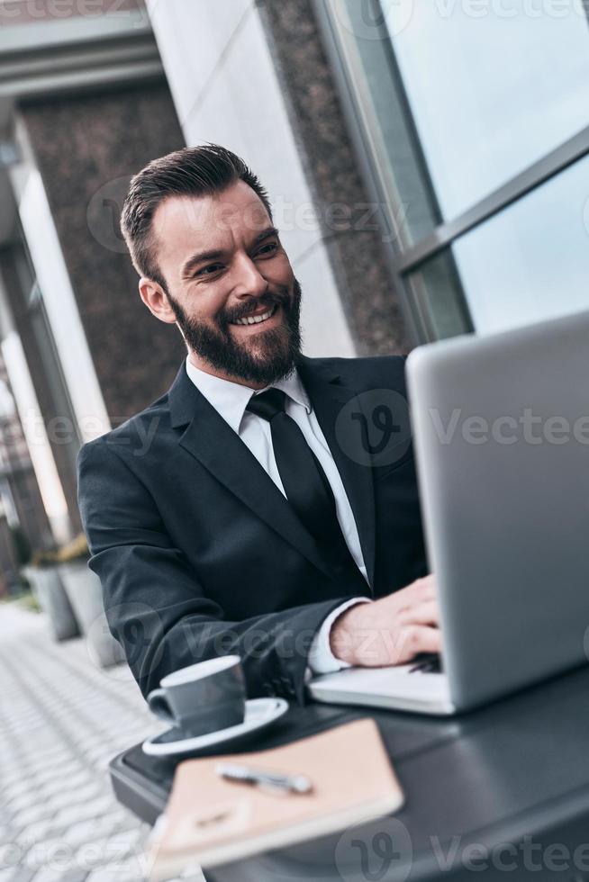 Achieving great results. Good looking young man in full suit using computer and smiling while sitting in the cafe outdoors photo