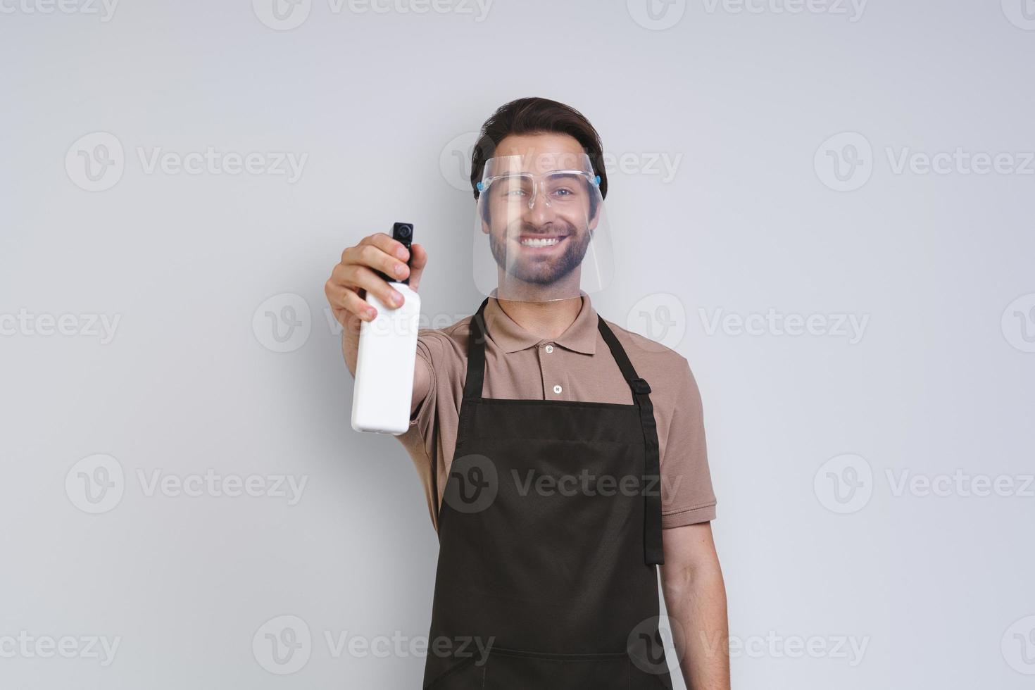 hombre joven guapo en delantal con protector facial y sosteniendo spray de limpieza mientras está de pie contra el fondo gris foto