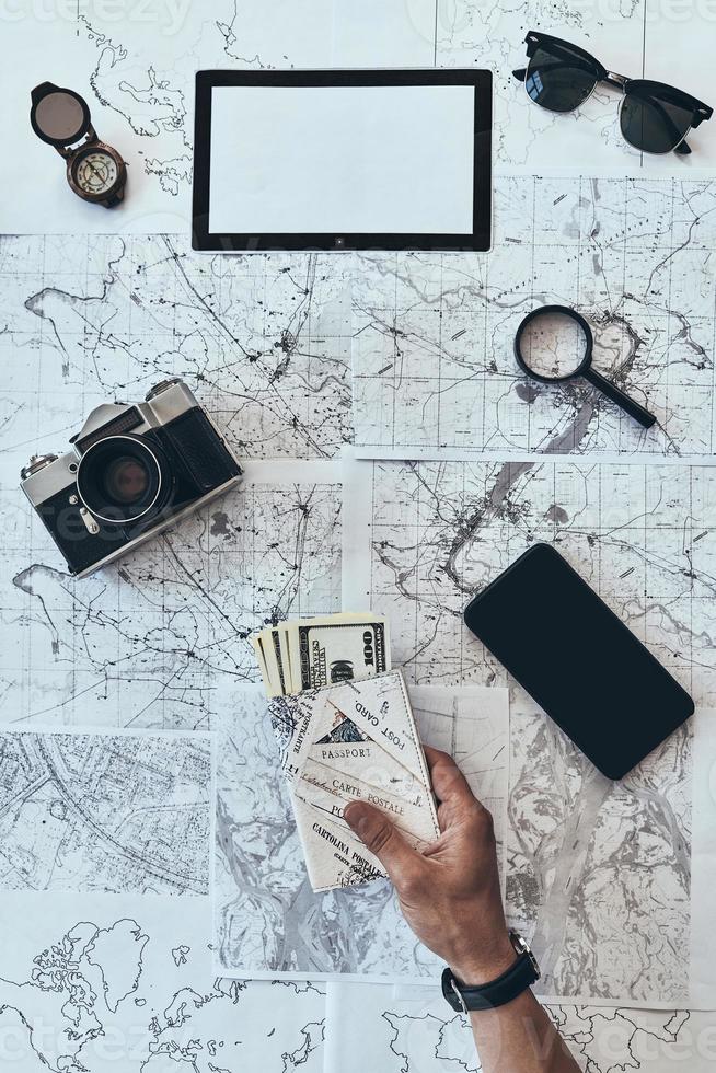 Preparing to travel. Close up top view of man holding passport and money with smart phone, sunglasses, photo camera, compass, magnifying glass lying on map around