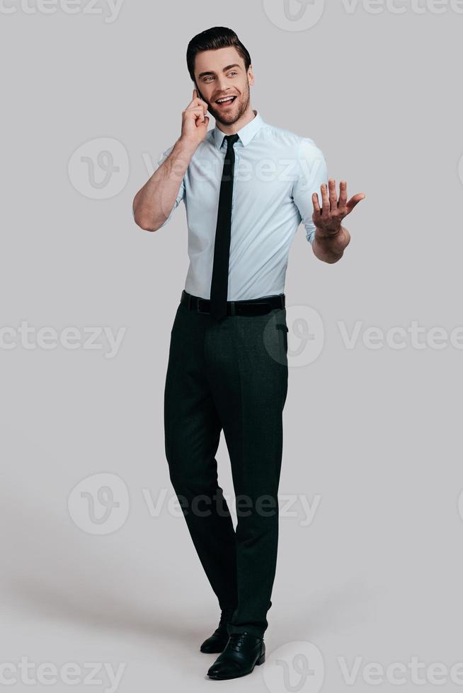 Sharing business ideas. Full length of handsome young man talking on smart phone and gesturing while standing against grey background photo