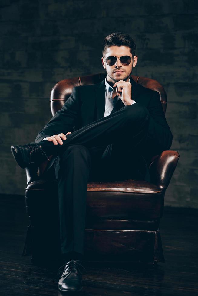 Mr. Perfection.Young handsome man in suit and sunglasses holding hand on chin and looking at camera while sitting in leather chair against dark grey background photo