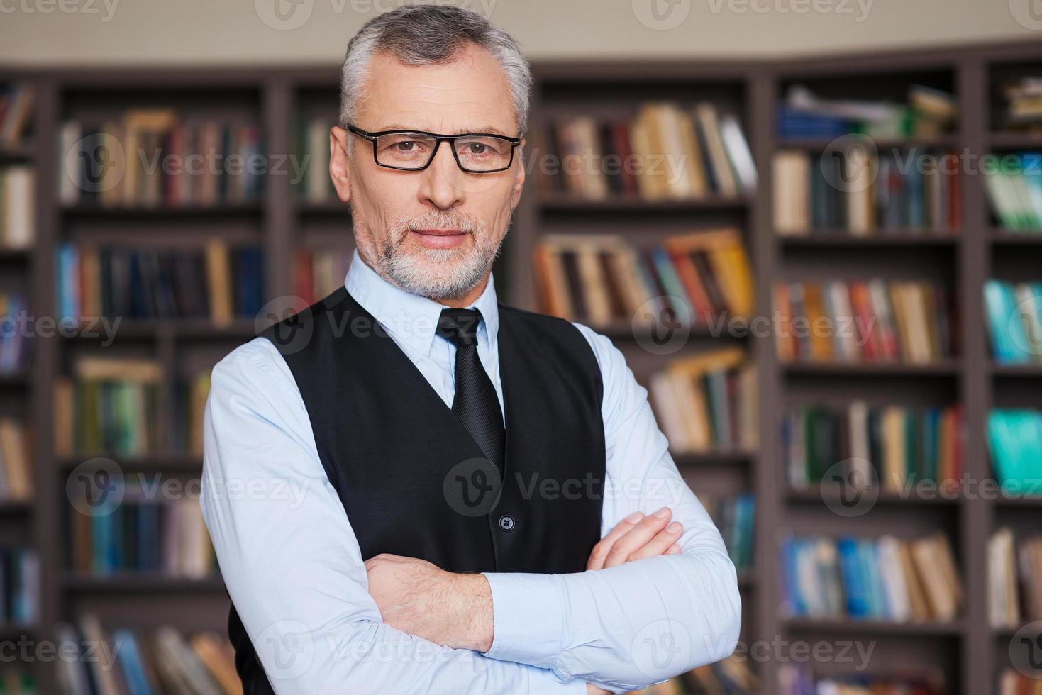 Confident and intelligence. Confident grey hair senior man in formalwear keeping arms crossed and looking at camera while standing against bookshelf photo