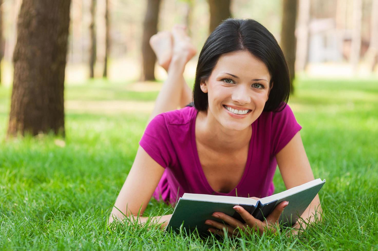 Beautiful bookworm in nature. Beautiful young woman reading book and smiling while lying in grass photo