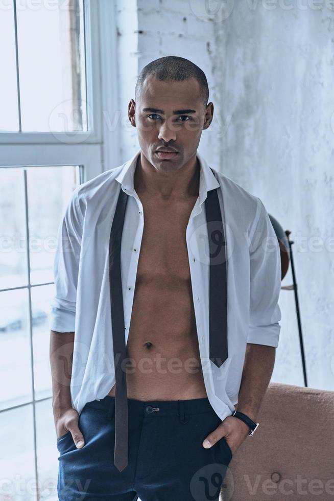 Real man. Handsome young African man in fully unbuttoned white shirt looking at camera while standing at home photo