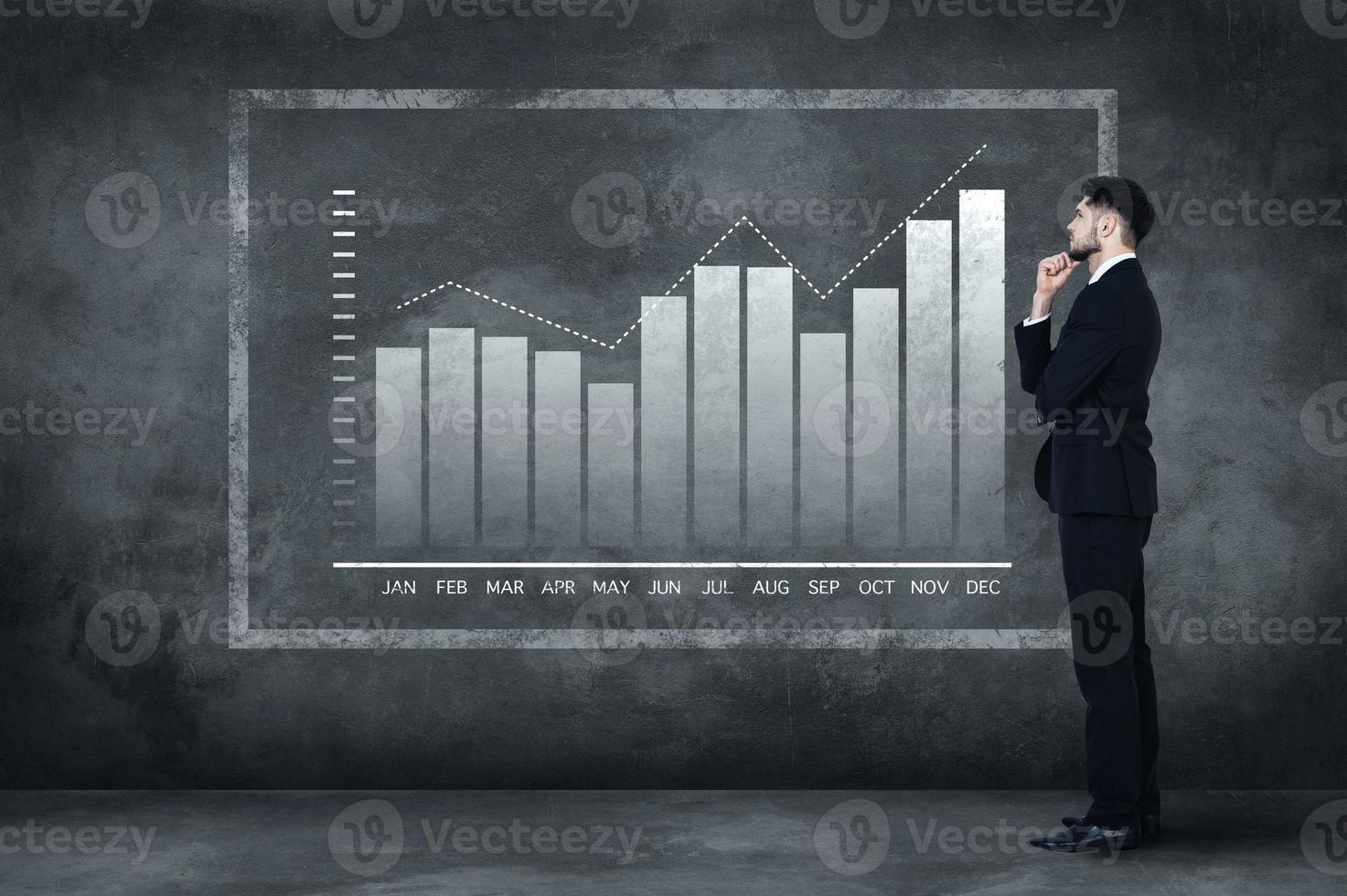 Thinking about solution. Full length of thoughtful young man in full suit standing near concrete wall and looking at the illustrated chart photo