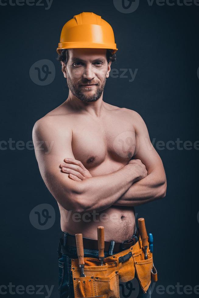 Muscular construction worker. Young confident man keeping arms crossed and looking at camera while standing against black background photo