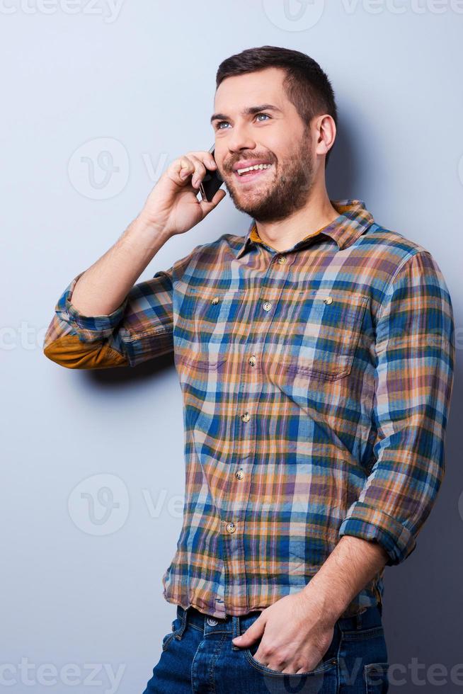 Communication with pleasure. Handsome young man in shirt holding mobile phone and looking away while standing against grey background photo