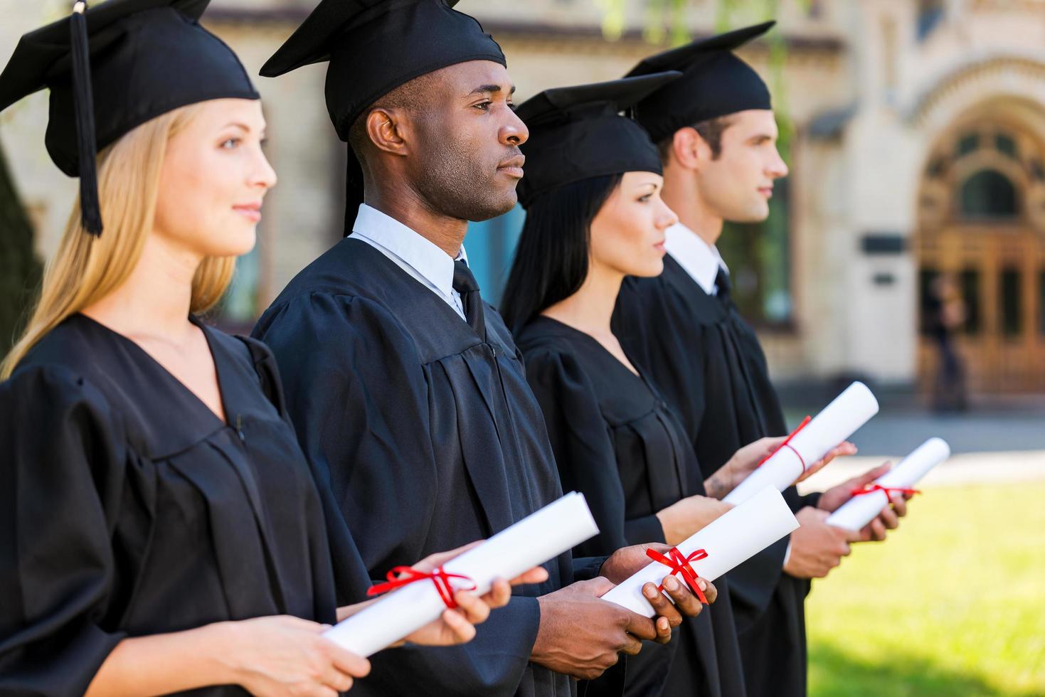 Looking to a bright future. Four college graduates holding their diplomas and looking away while standing in a row and in front of the university photo