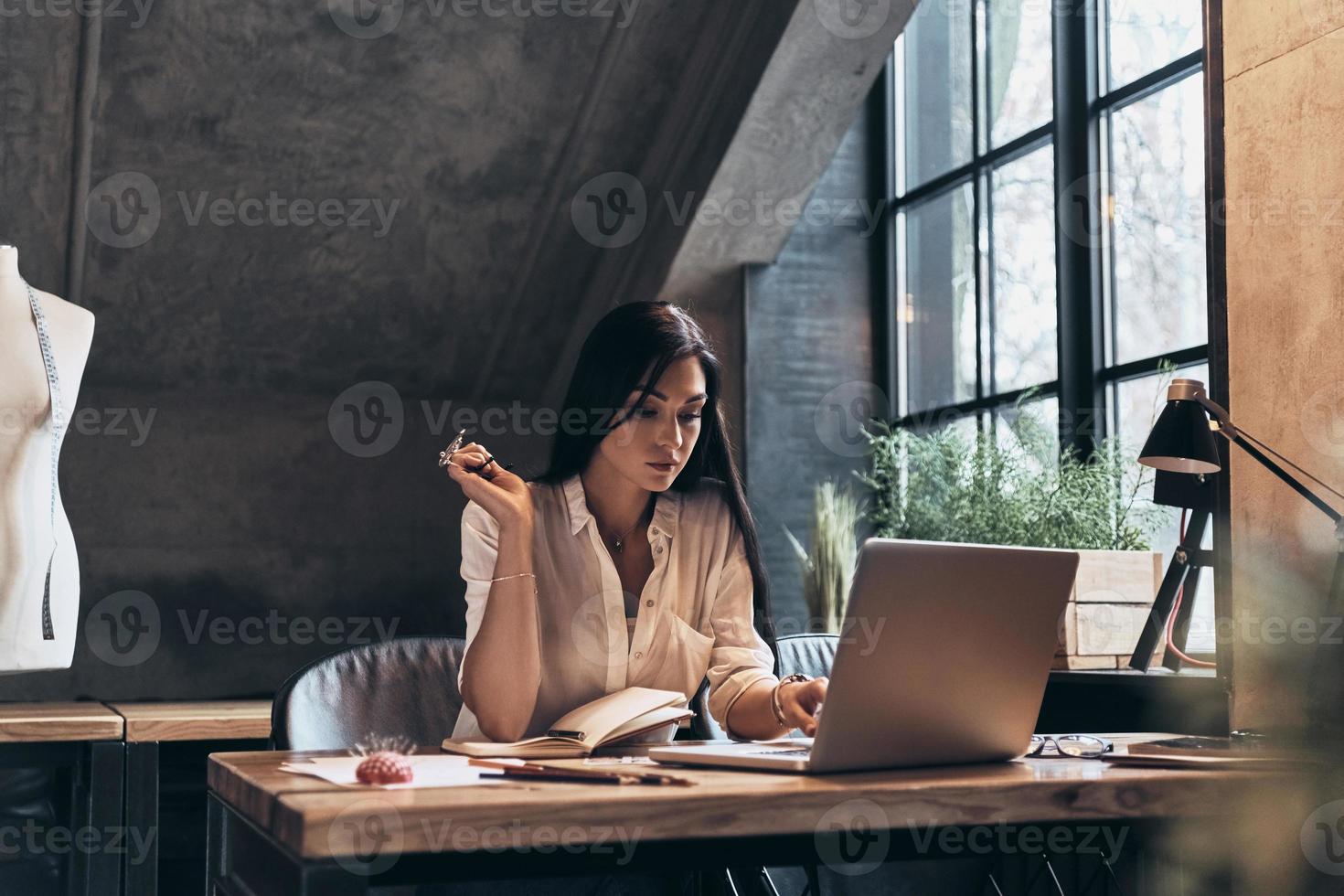 Developing  project. Concentrated young woman working using laptop while sitting in her workshop photo