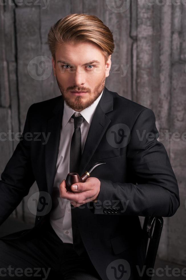 Man with a smoking pipe. Handsome young man in formalwear holding a smoking pipe and looking at camera while sitting on the chair photo