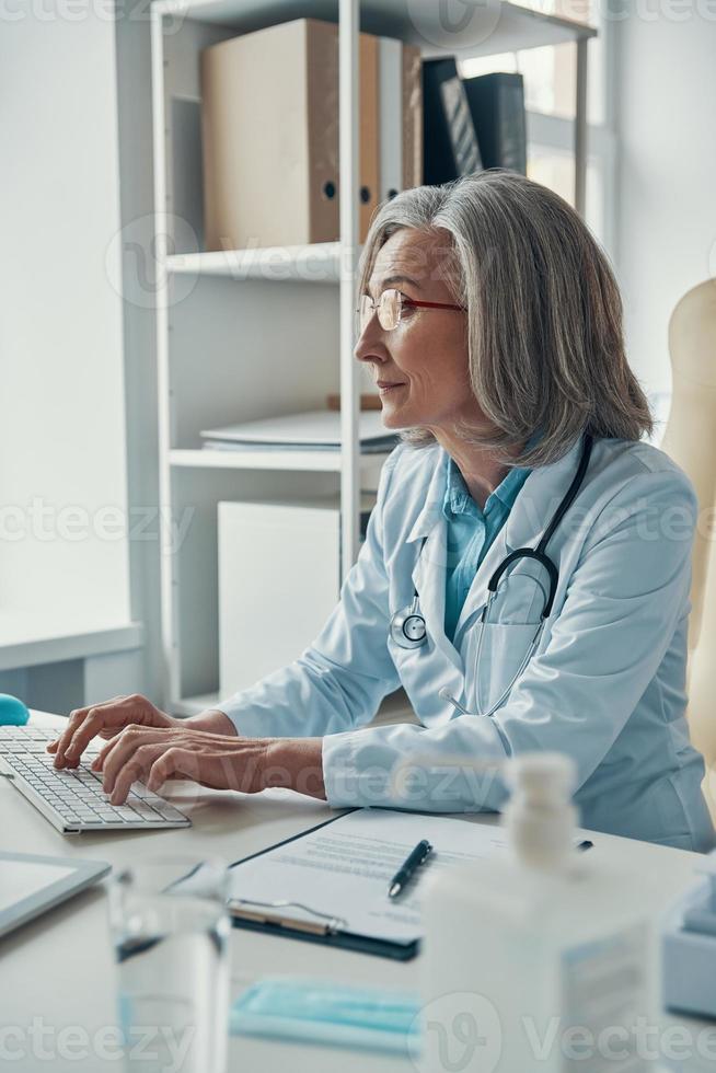 Serious mature female doctor in white lab coat working using computer while sitting in her office photo