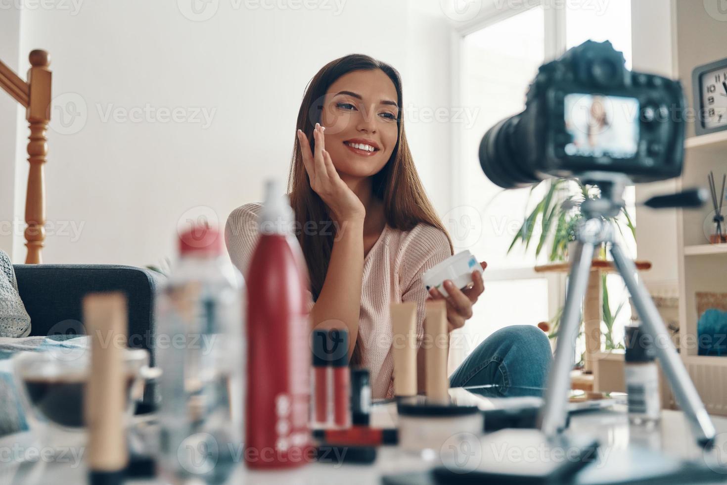 Beautiful young woman applying lip gloss and smiling while making social media video photo