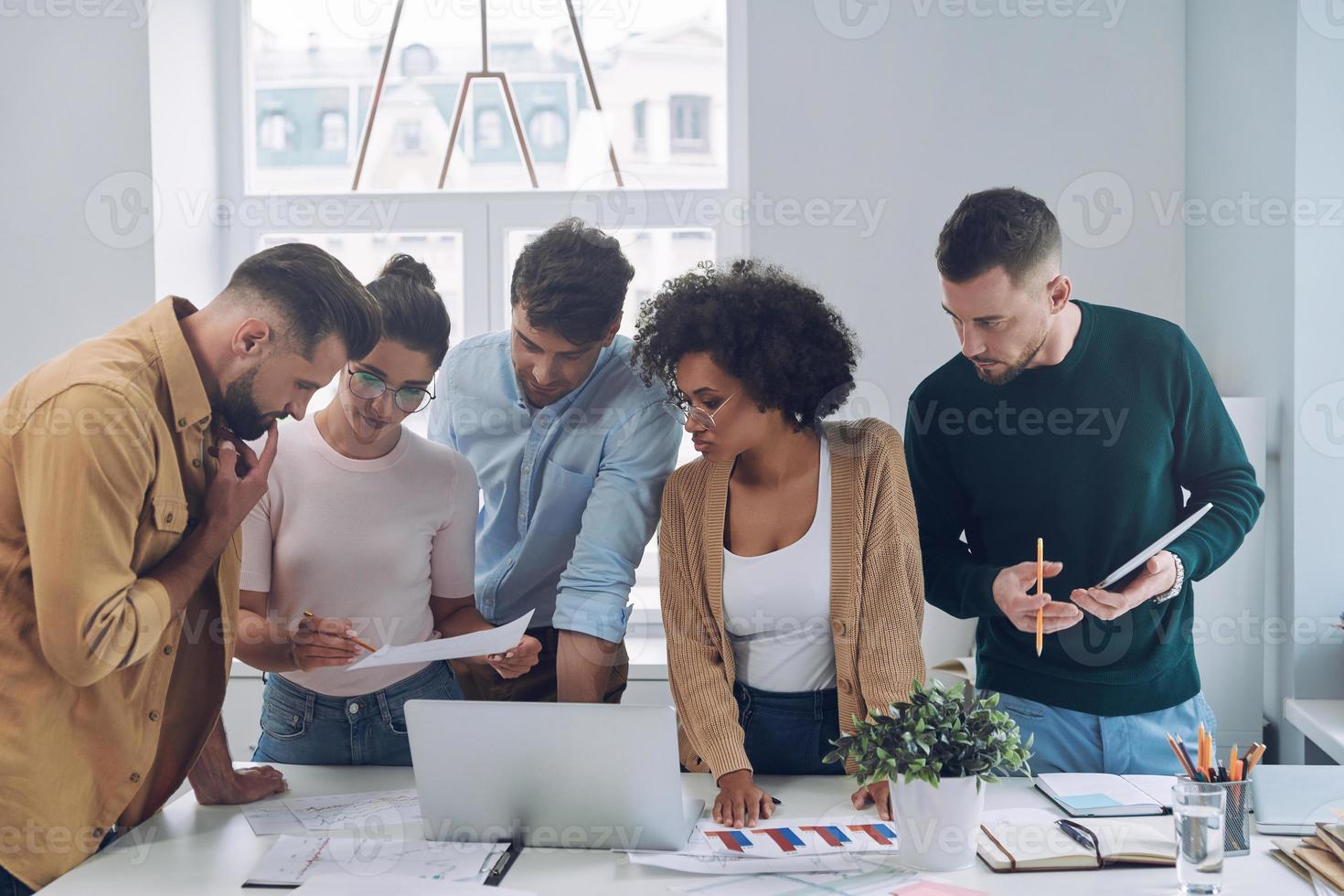 grupo de jóvenes confiados en ropa casual inteligente discutiendo negocios mientras están de pie cerca del escritorio en la oficina foto