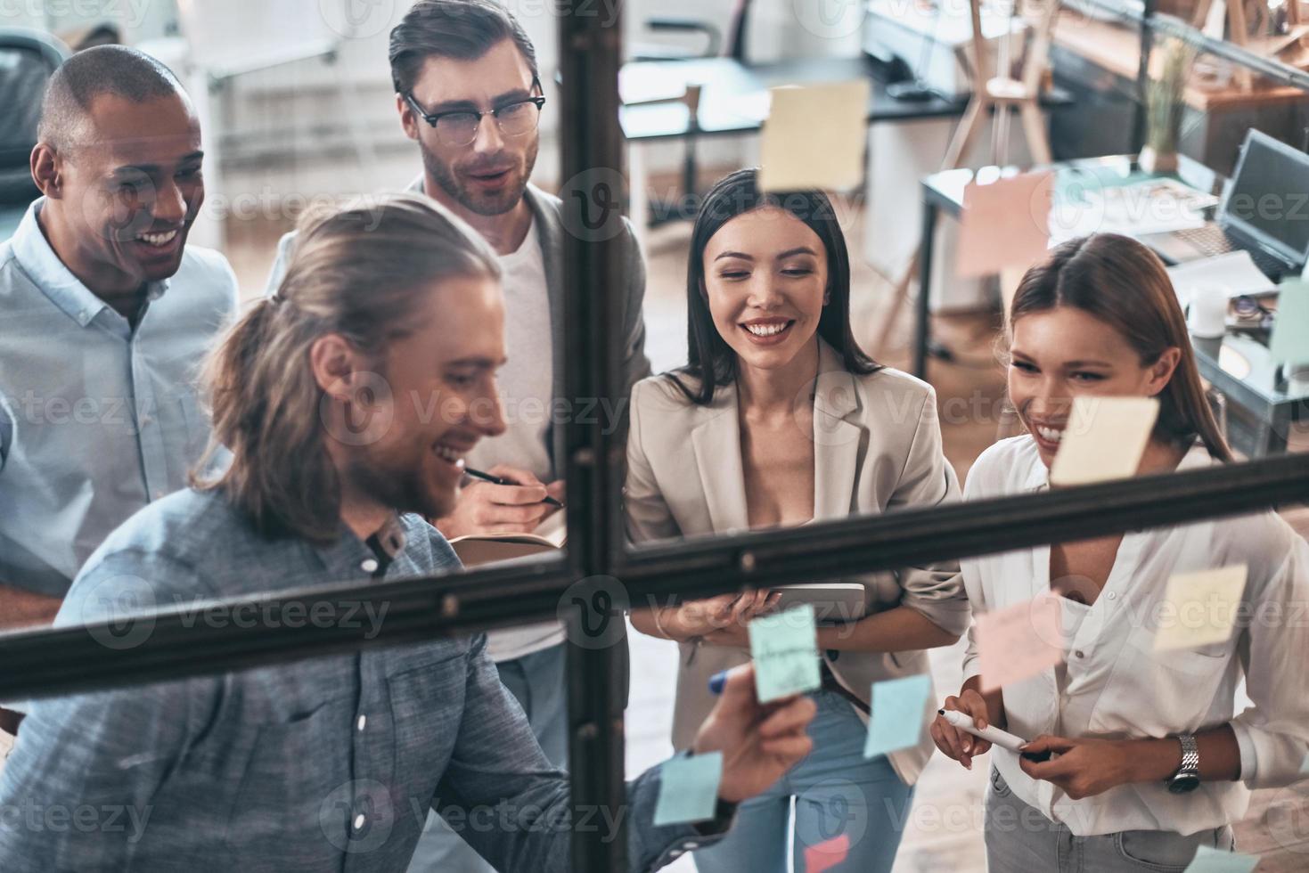 Ensuring success by working as a team. Top view of young modern people in smart casual wear using adhesive notes while standing behind the glass wall in the board room photo