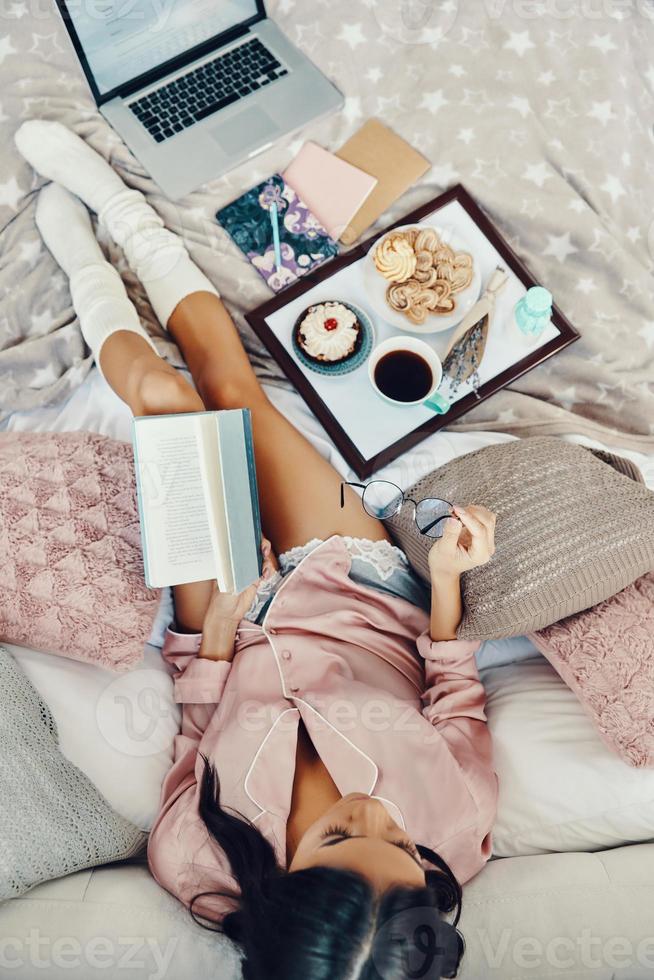 Top view of beautiful young woman in pajamas holding notepad and eyewear while resting in bed at home photo