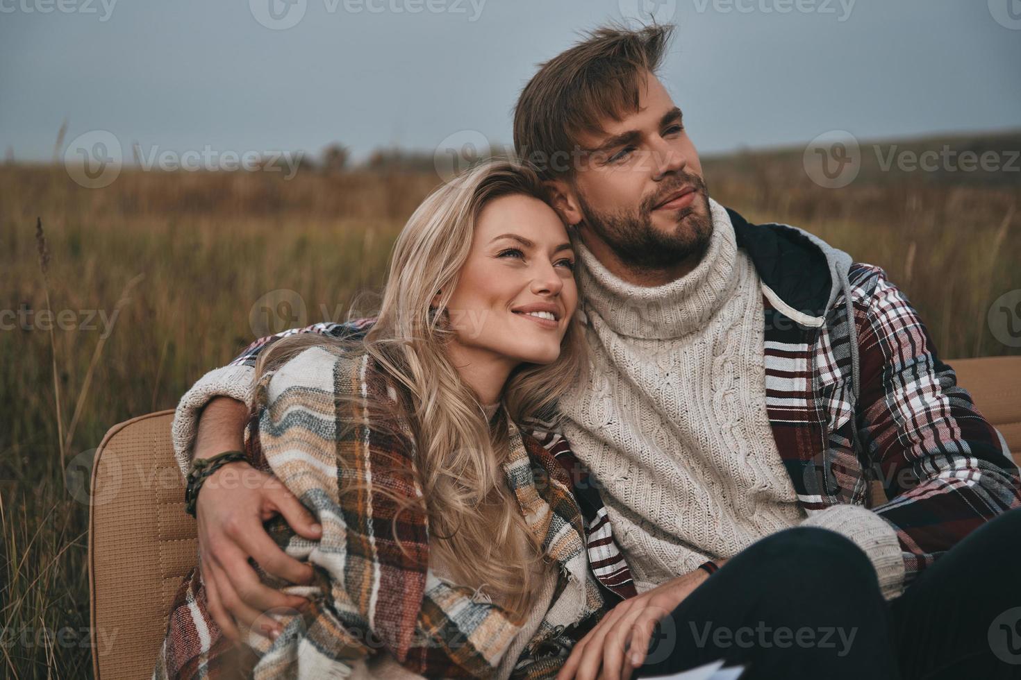 todas las preocupaciones se fueron volando... hermosa pareja joven abrazándose y mirando hacia otro lado con una sonrisa mientras se sienta en el campo foto