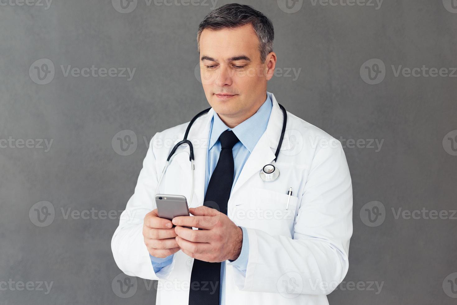 Looking through his timetable. Mature male doctor using his smart phone while standing against grey background photo