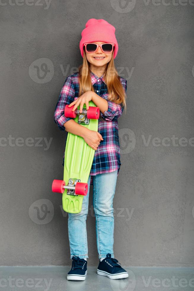 genial chica patinadora. longitud completa de una niñita alegre con gafas de sol mirando a la cámara con una sonrisa y sosteniendo una patineta mientras está de pie contra un fondo gris foto