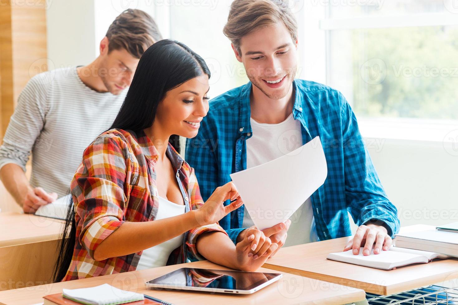 aquí hay información que necesitamos. un joven sonriente sosteniendo un papel mientras una mujer lo señalaba y se sentaba cerca de él en el escritorio del salón de clases foto