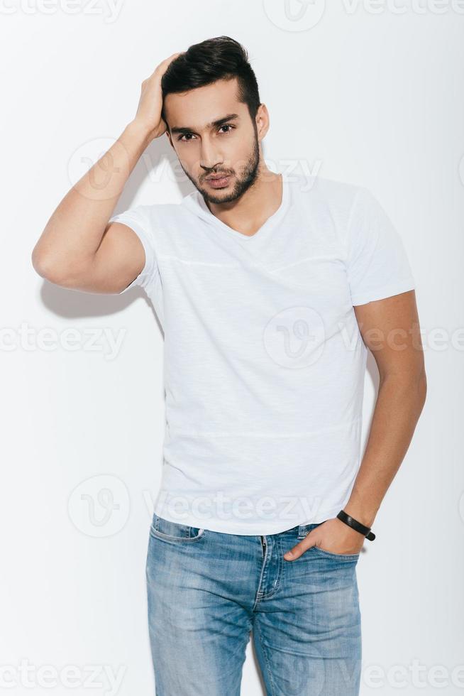 Used to look good. Handsome young Indian man holding hand in hair and looking at camera while standing against white background photo