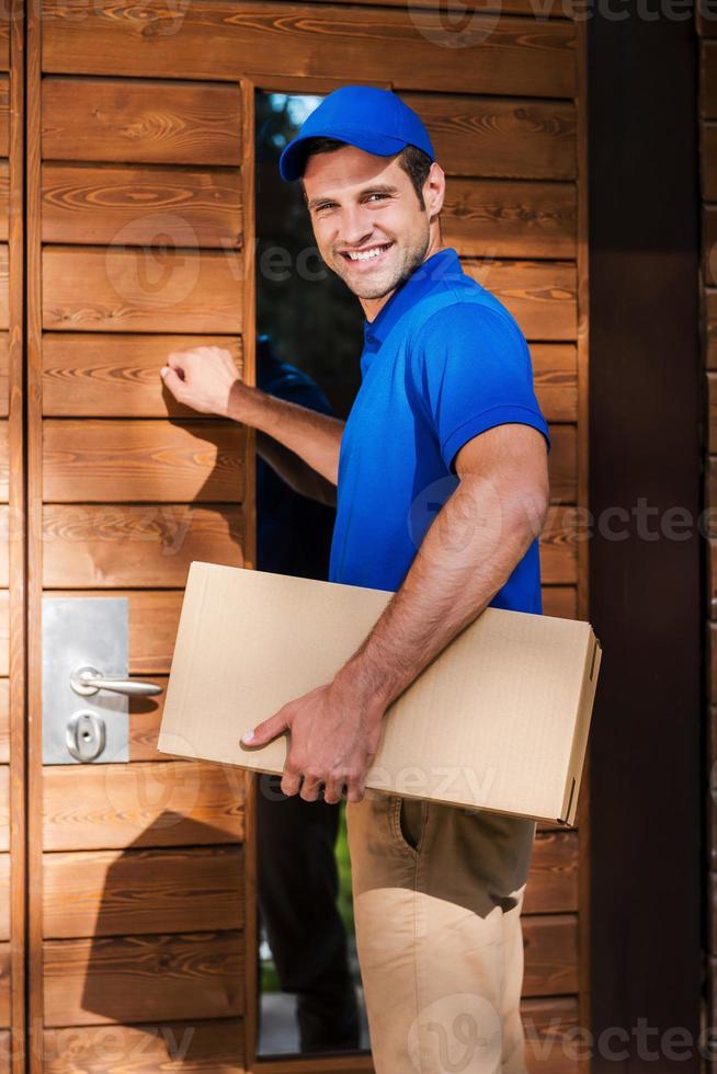 entrega a tu casa. joven mensajero sonriente sosteniendo una caja de cartón mientras llama a la puerta de una casa residencial foto