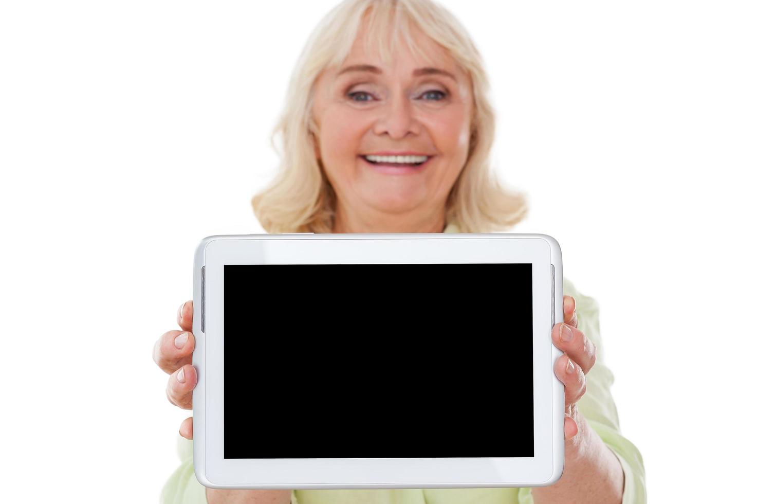 Copy space on her tablet. Cheerful senior woman stretching out digital tablet and smiling while standing isolated on white background photo