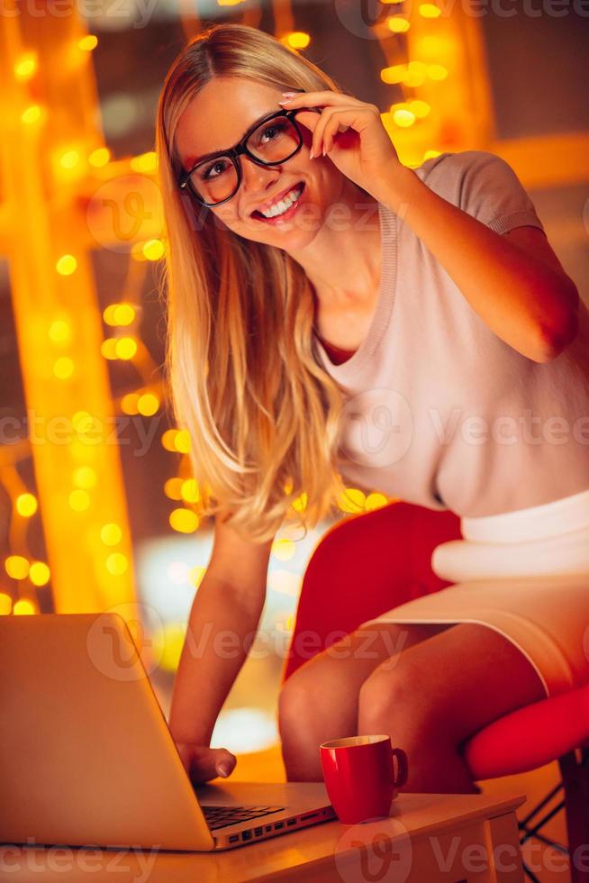 Enjoying work even late hours. Beautiful young woman in smart casual wear looking t camera and smiling while working on laptop and with Christmas lights in the background photo