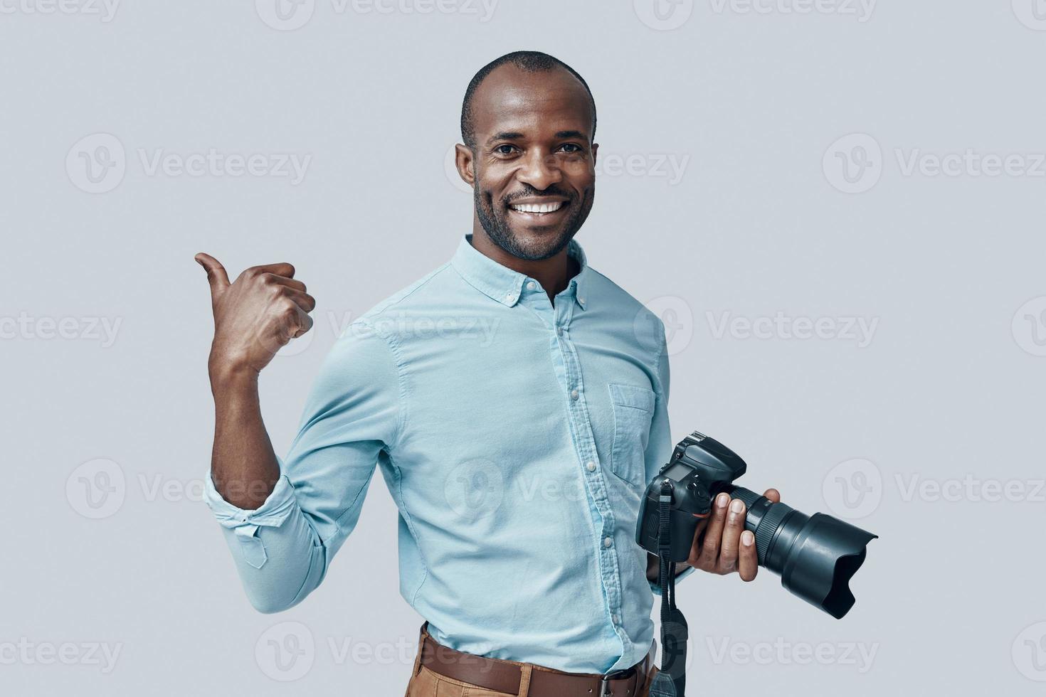 Happy young African man using digital camera and smiling while standing against grey background photo