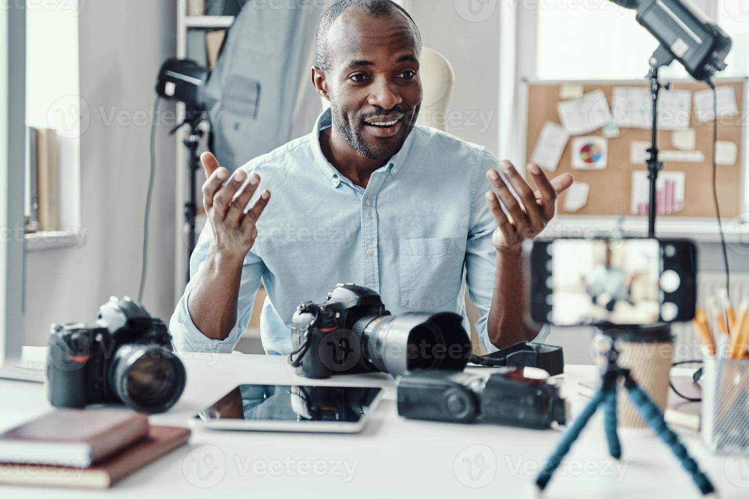 Charming young African man in shirt showing digital camera and telling something while making social media video photo