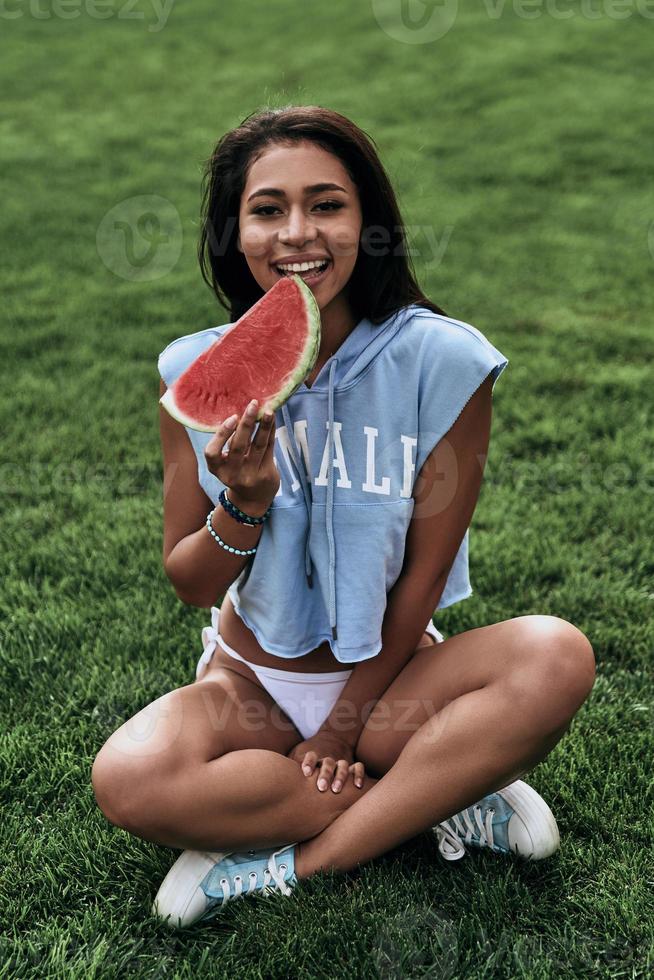 disfrutando de un refrigerio saludable. mujer joven atractiva comiendo una rebanada de sandía y sonriendo mientras se sienta en la hierba foto