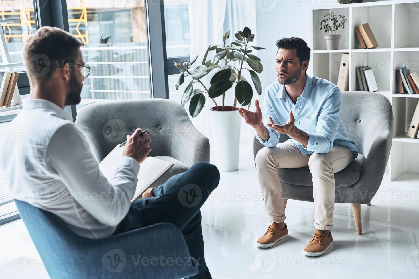 Trying to help. Young frustrated man speaking and gesturing while sitting on the therapy session with psychologist photo