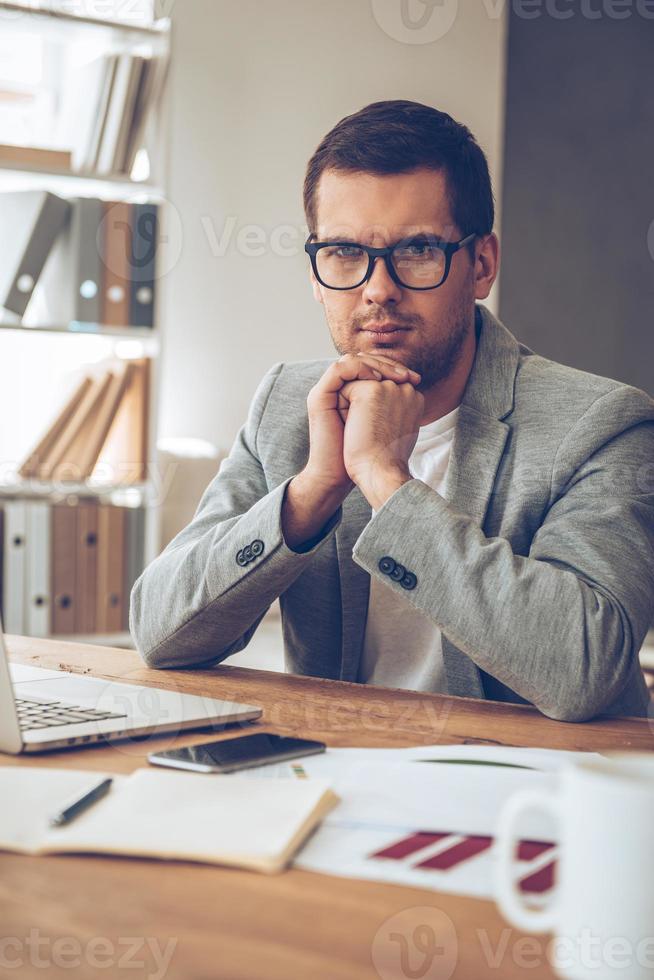 Making business every day. Handsome pensive young man in glasses looking at camera while sitting at his working place photo