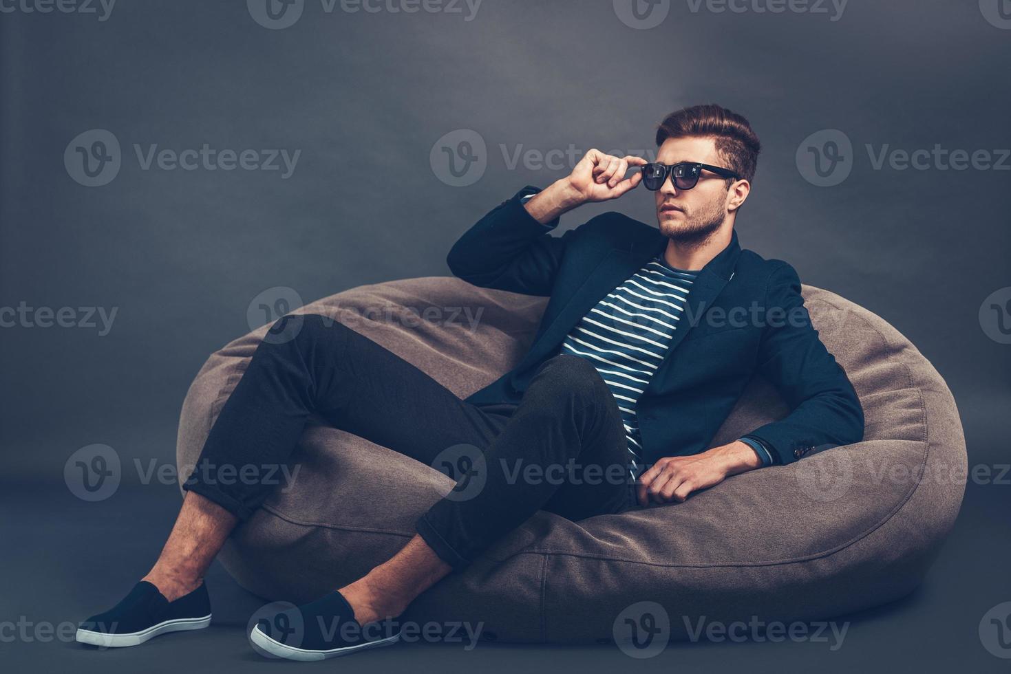 guapo y elegante. un joven apuesto y seguro de sí mismo ajustando sus gafas de sol y mirando hacia otro lado mientras se sienta en una bolsa de frijoles contra un fondo gris foto