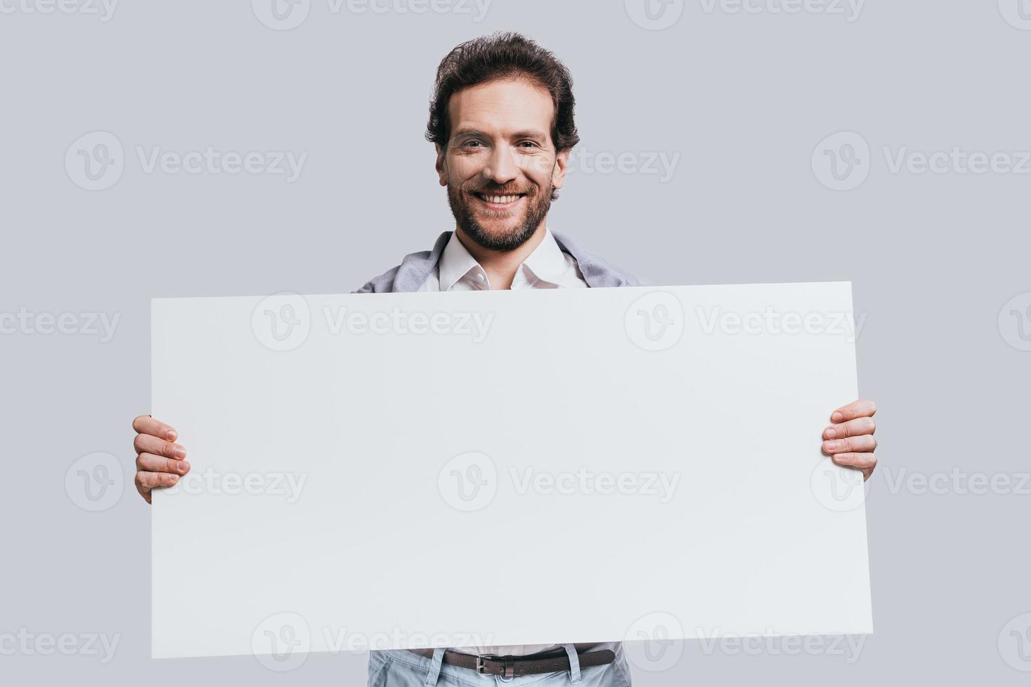 llevando espacio de copia. joven confiado vestido con ropa informal elegante que sostiene un rotafolio en blanco y sonríe mientras se enfrenta a un fondo gris foto